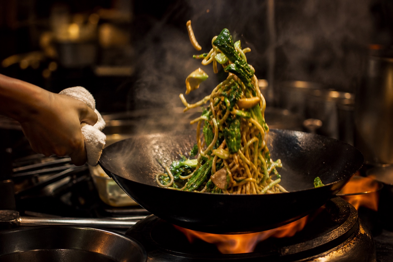 A chef tossing hand-pulled noodles in a wok over open flame